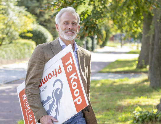 Léo Pfrommer, makelaar van Pfrommer Makelaardij met een Te Koop-bord onder de arm in een groene laan